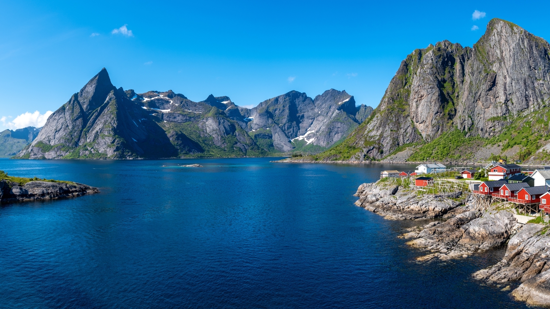 A picturesque view of red cabins perched on the rocky shore of a fjord in Norway, surrounded by majestic mountains and a clear blue sky.
