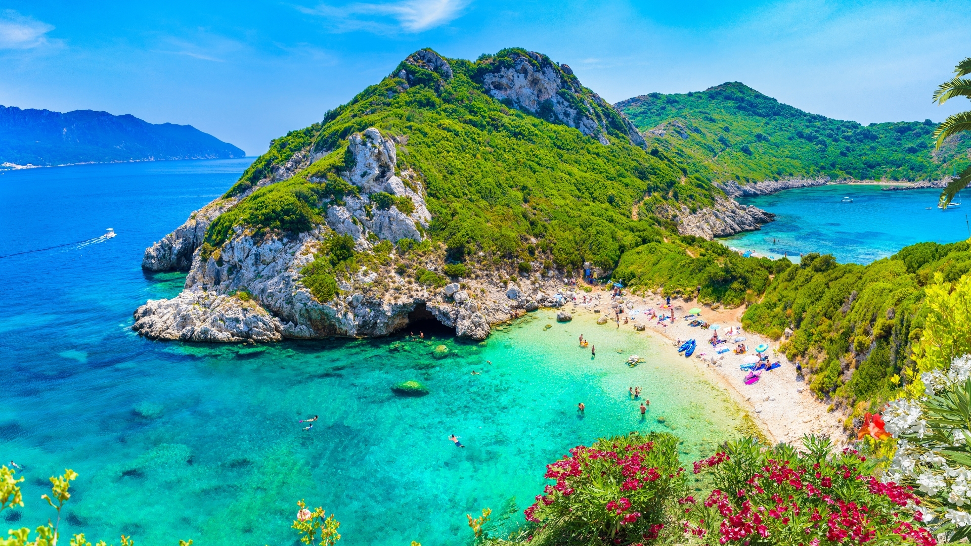 The image shows a stunning aerial view of Porto Timoni in the Afionas region of Corfu, featuring a beautiful beach with clear turquoise waters, surrounded by lush green hills and rocky cliffs. People can be seen enjoying the beach, and there are boats in the water, creating a picturesque coastal scene.