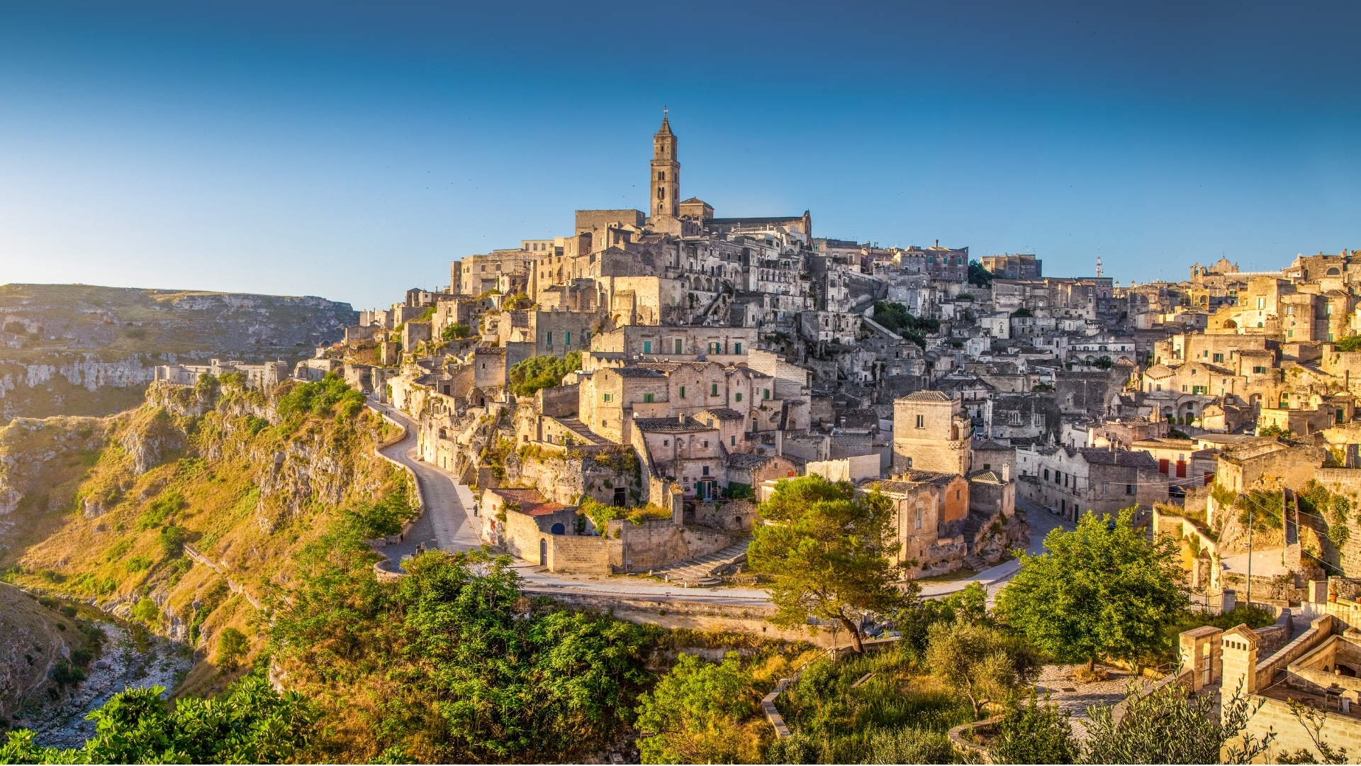 The image depicts the ancient town of Matera in Basilicata, Italy, at sunrise. The town is characterized by its unique stone buildings and structures that are built into the rocky landscape, with a prominent bell tower visible in the background. The scene captures the warm glow of the early morning light illuminating the historic architecture and the surrounding greenery.