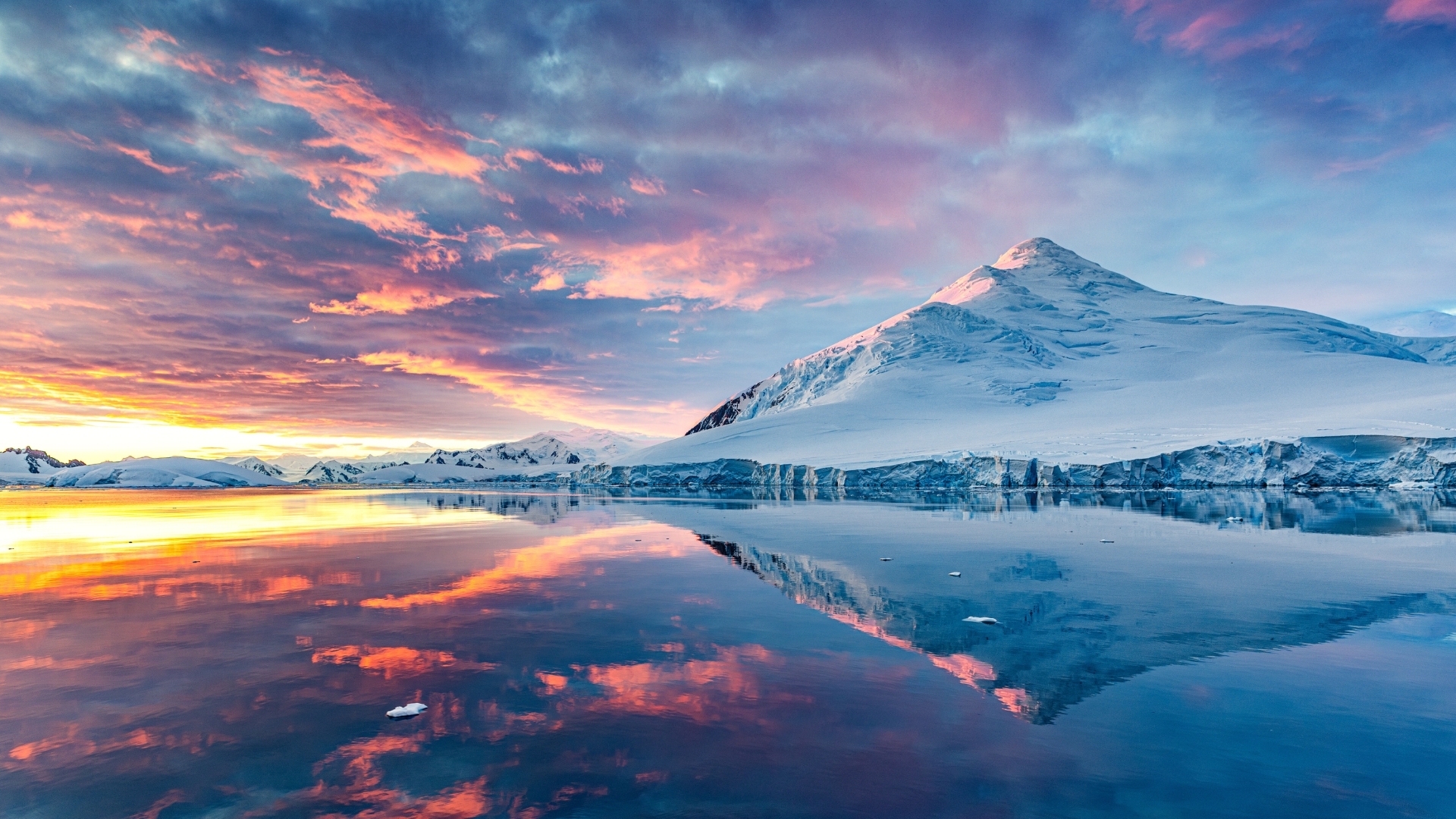A stunning landscape of Antarctica featuring majestic mountains next to a calm sea, reflecting the vibrant colors of the sky during the midnight sun.