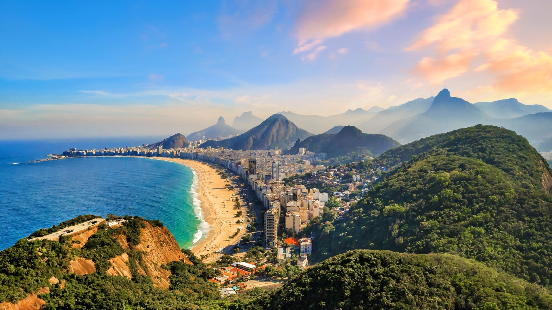 The image shows a panoramic view of Copacabana Beach and Ipanema Beach in Rio de Janeiro, Brazil, with lush green mountains in the foreground and a clear blue sky above. The coastline features sandy beaches and a bustling cityscape, highlighting the natural beauty and urban life of the area.