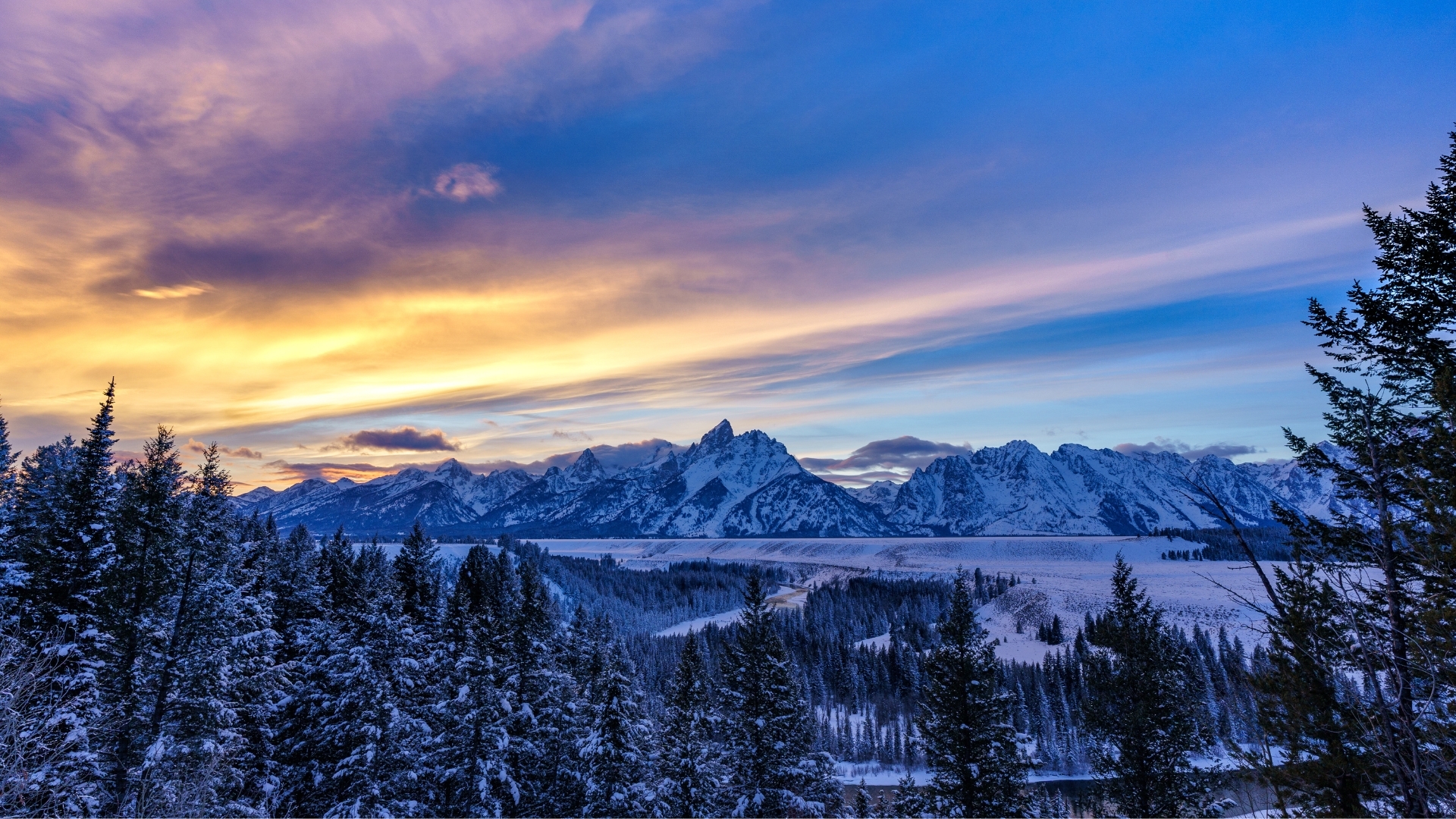 The image depicts a dramatic sunset over the Grand Tetons in Jackson Hole during winter, showcasing snow-covered mountains under a colorful sky with shades of orange, purple, and blue, framed by evergreen trees in the foreground.
