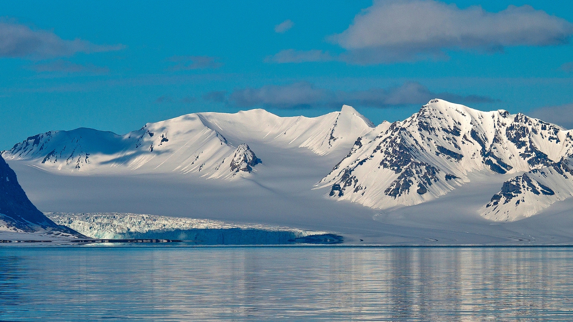 The image depicts a stunning landscape of glacier and snowcapped mountains in Oscar II Land, Arctic. The scene features majestic peaks covered in white snow, reflecting in the calm waters below, under a clear blue sky with a few clouds.