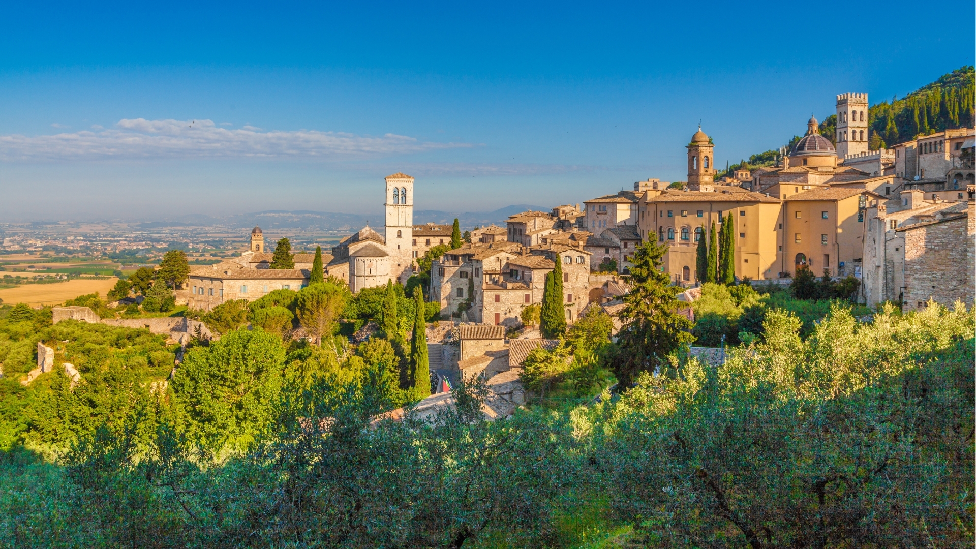The image depicts the historic town of Assisi in Umbria, Italy, bathed in the warm light of sunrise. The scene showcases a picturesque landscape with medieval stone buildings, a bell tower, and lush greenery, set against a clear blue sky and rolling hills in the background.
