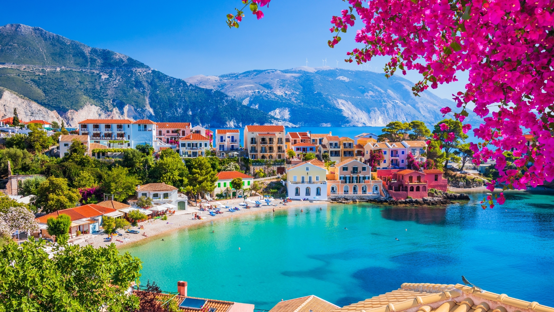The image depicts the colorful village of Assos in Kefalonia, Greece, showcasing vibrant buildings along a serene bay with clear turquoise waters, surrounded by lush greenery and mountains. Bougainvillea flowers add a splash of pink to the scene, enhancing the picturesque coastal view.