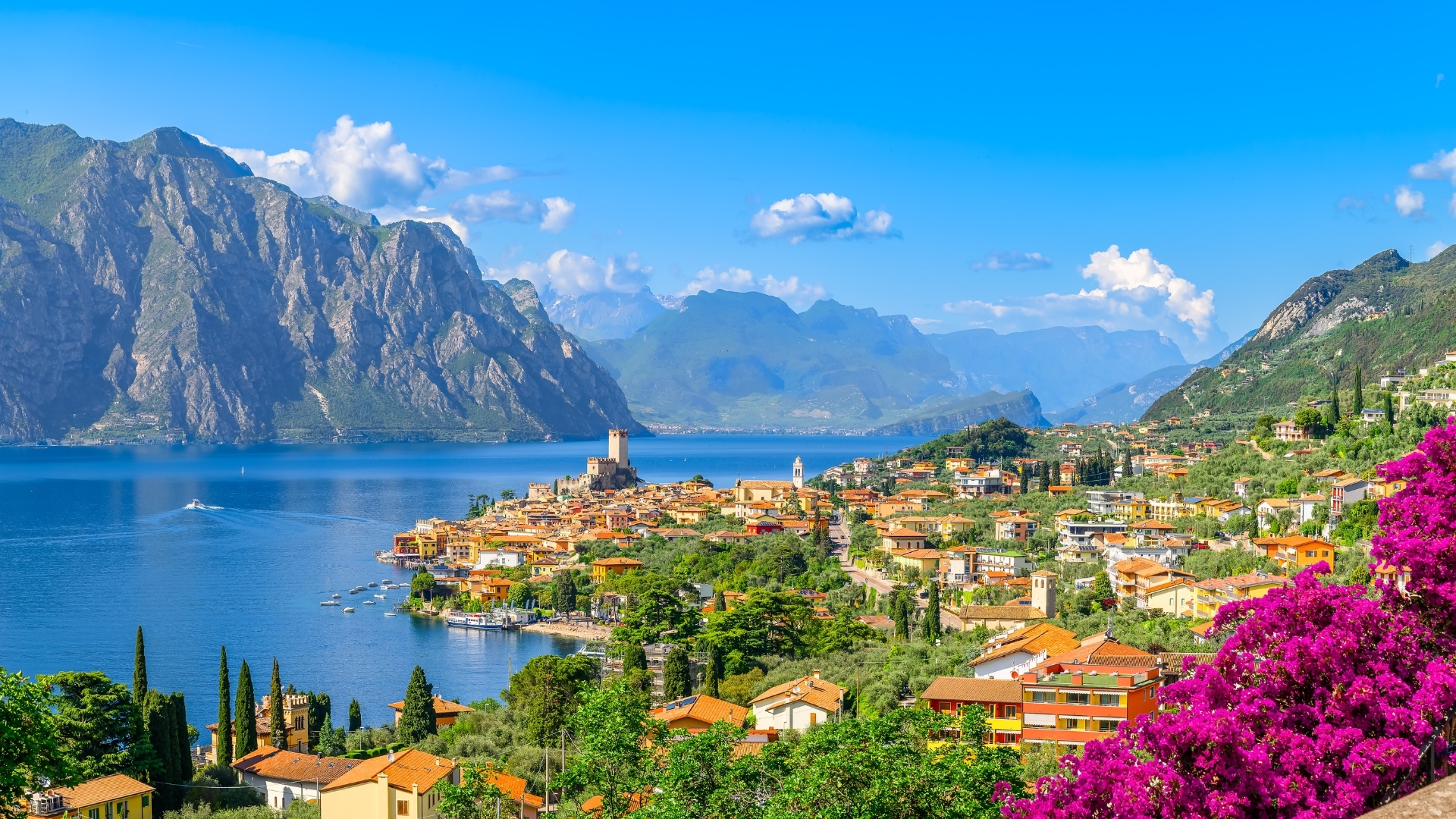 The image depicts a picturesque view of Malcesine, a charming town located on the shores of Lake Garda in Italy. The scene features colorful buildings nestled among lush greenery, with a backdrop of majestic mountains and a clear blue sky. A few boats can be seen on the tranquil lake, enhancing the serene atmosphere of this beautiful landscape.