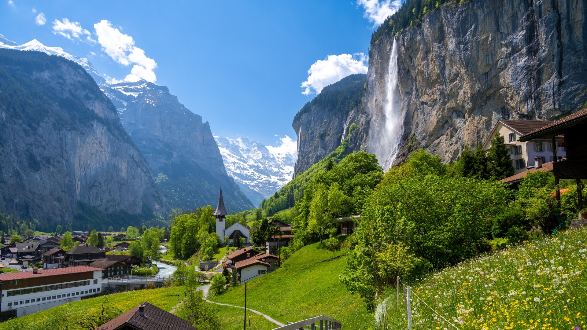 The image depicts the picturesque village of Lauterbrunnen in Switzerland, featuring a charming church and a stunning waterfall cascading down a rocky cliff, surrounded by lush greenery and majestic mountains under a clear blue sky.