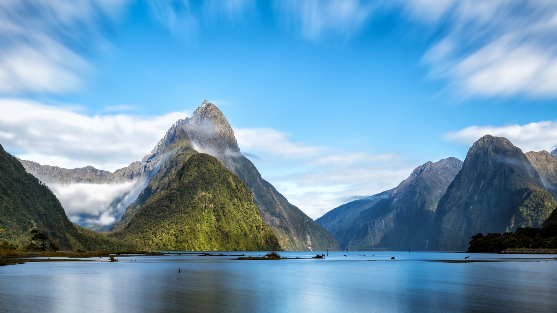 The image depicts Milford Sound in New Zealand, showcasing towering mountains with lush greenery, a serene body of water, and a bright blue sky with wispy clouds. The landscape is characterized by dramatic peaks and a tranquil atmosphere, highlighting the natural beauty of this iconic location.