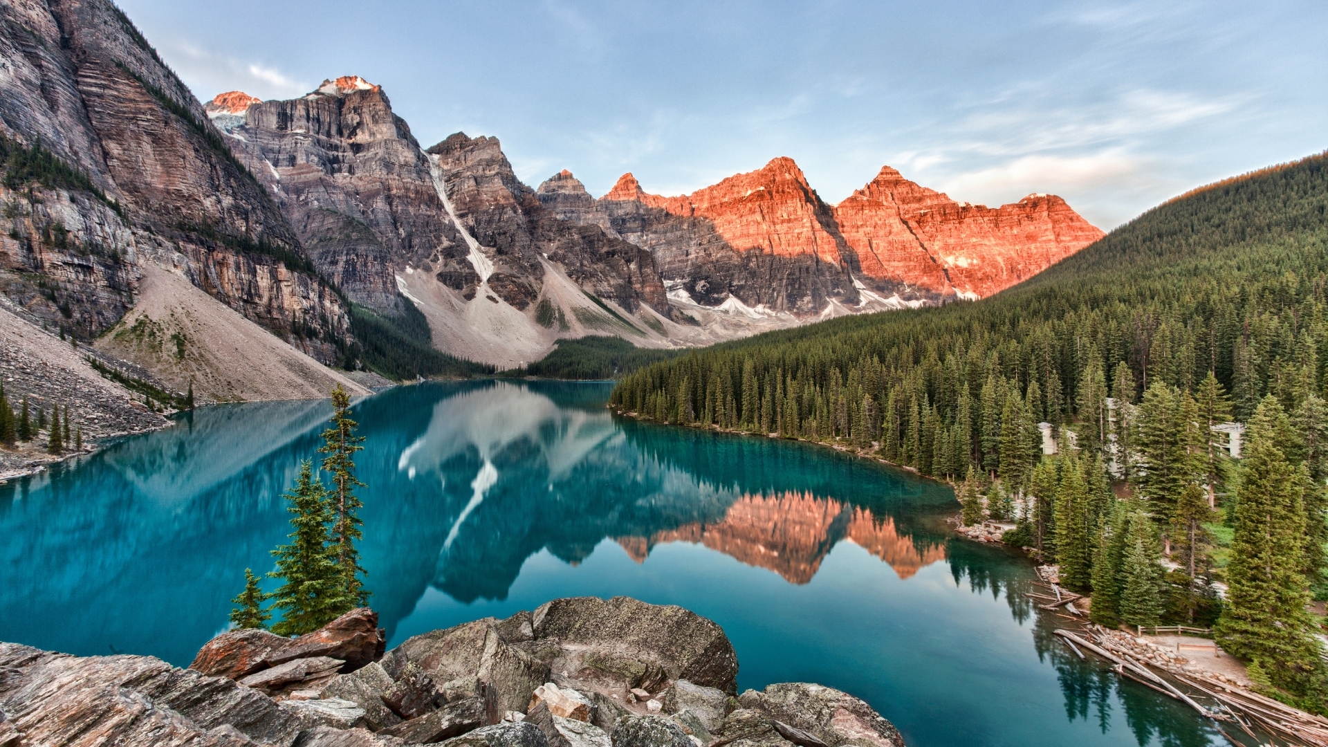 The image depicts Moraine Lake in Banff National Park, Canada, showcasing stunning turquoise waters reflecting the surrounding mountains at sunrise, with vibrant colors illuminating the peaks and lush green forests lining the shore.