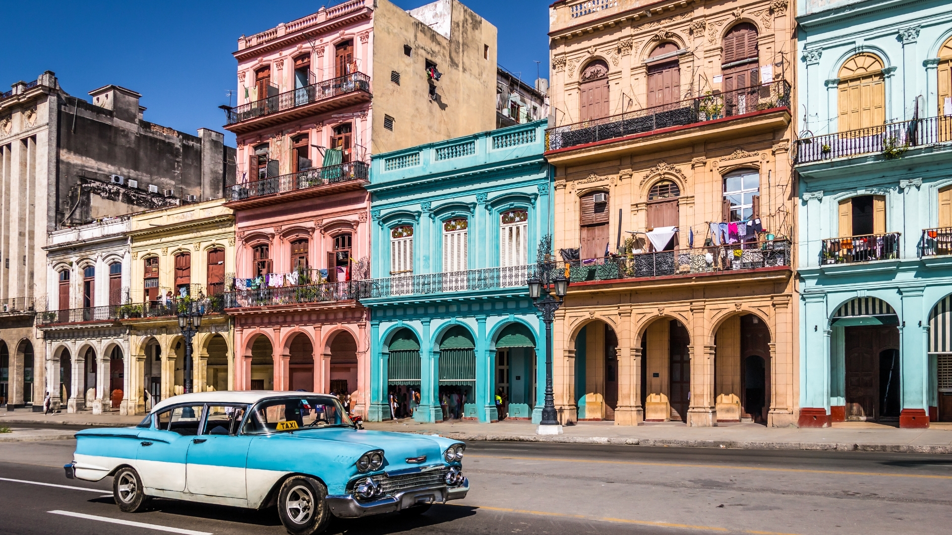 The image depicts a vibrant street scene in Old Havana, Cuba, showcasing colorful colonial buildings in shades of pink, yellow, blue, and beige. A classic blue and white vintage car, likely a taxi, is seen driving along the street, while laundry hangs from balconies, adding to the lively atmosphere of the area.
