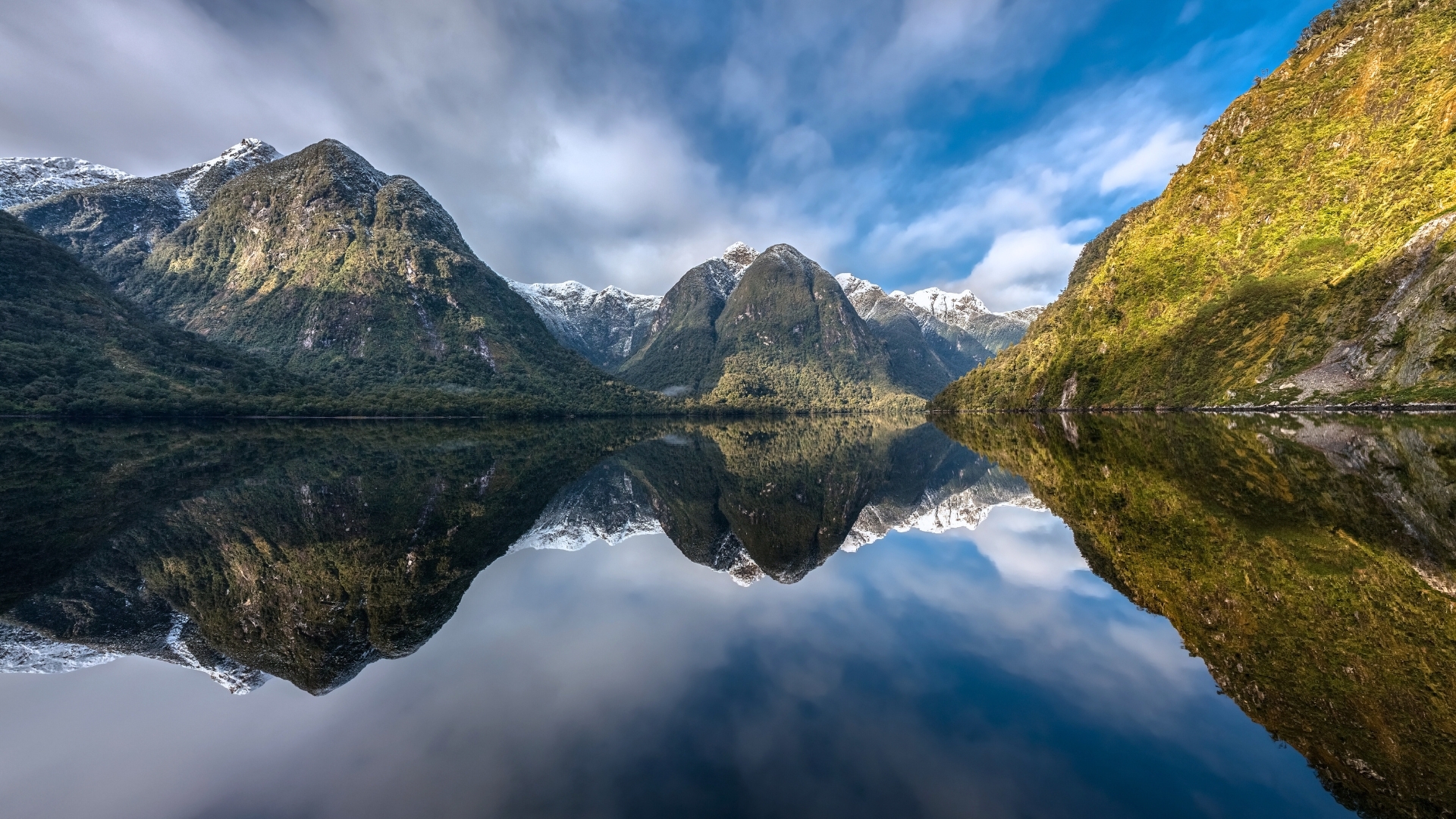 A panoramic view of Doubtful Sound in New Zealand, showcasing majestic mountains reflected in calm waters under a partly cloudy sky.