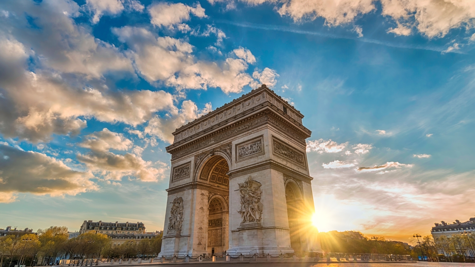 The image depicts the Arc de Triomphe in Paris during sunset, with a vibrant sky filled with clouds and the sun shining behind the monument, creating a beautiful silhouette effect.