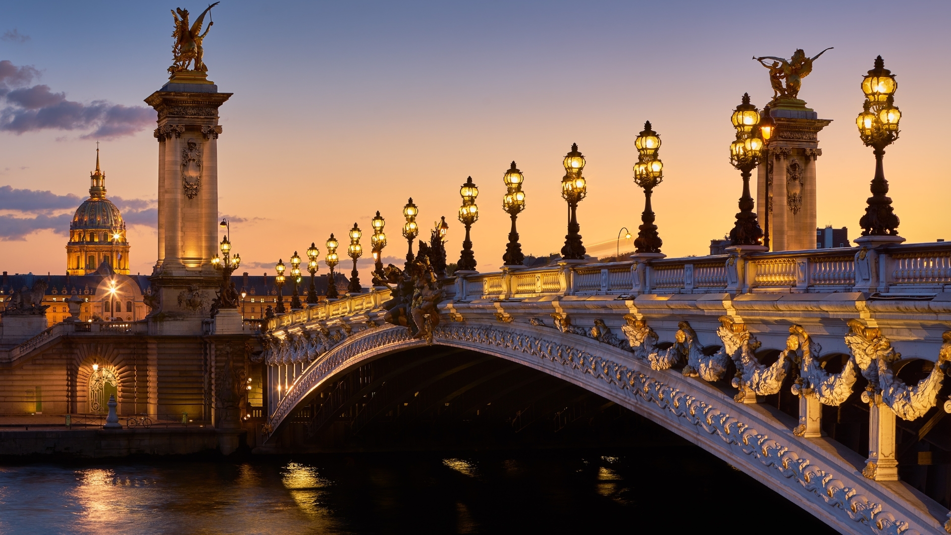The image depicts the Pont Alexandre III bridge in Paris, beautifully illuminated by lamp posts at sunset. The bridge features ornate sculptures and golden details, with the dome of Les Invalides visible in the background, creating a picturesque scene against the colorful sky.