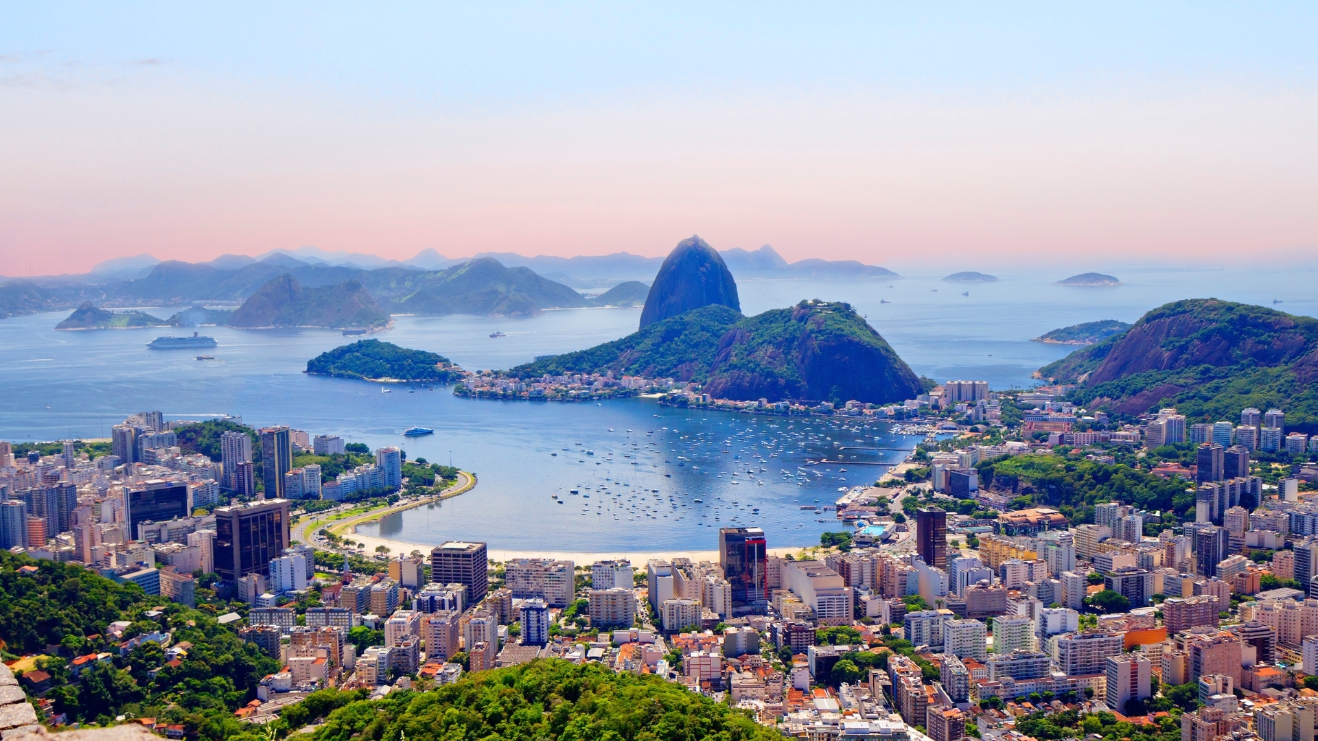 The image shows a panoramic view of Rio de Janeiro, Brazil, from the Corcovado mountain. It features the city's skyline, lush green hills, and the iconic Sugarloaf Mountain in the background, with a serene bay dotted with boats and islands.
