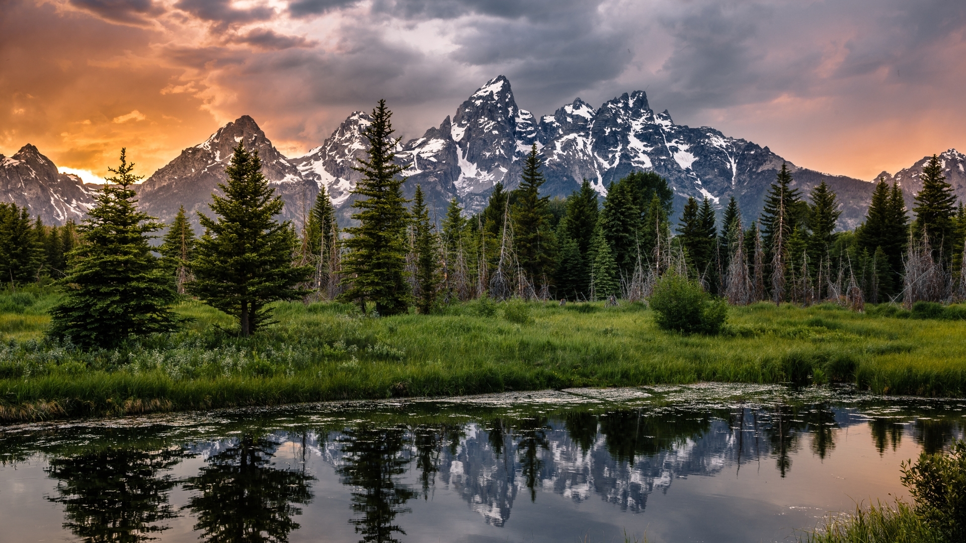 The image depicts a stunning sunset over the Grand Teton Range, with vibrant orange and purple hues in the sky. In the foreground, lush green trees and a calm body of water reflect the mountains and the colorful sky, creating a serene and picturesque landscape.