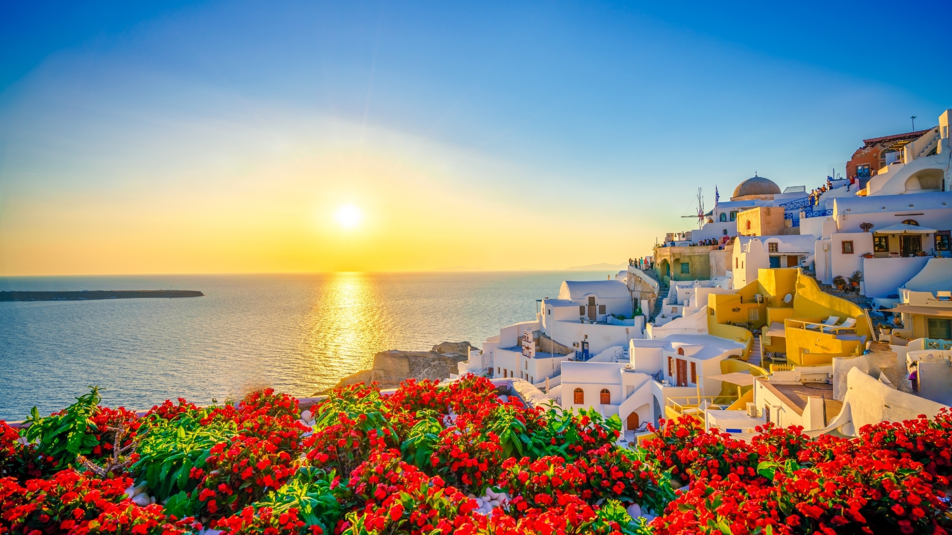 The image depicts a stunning sunset over the Aegean Sea in Oia, Santorini, Greece. The sky is painted in warm hues of orange and yellow, reflecting on the water, while vibrant red flowers in the foreground contrast beautifully with the white and colorful buildings of the town, creating a picturesque scene.
