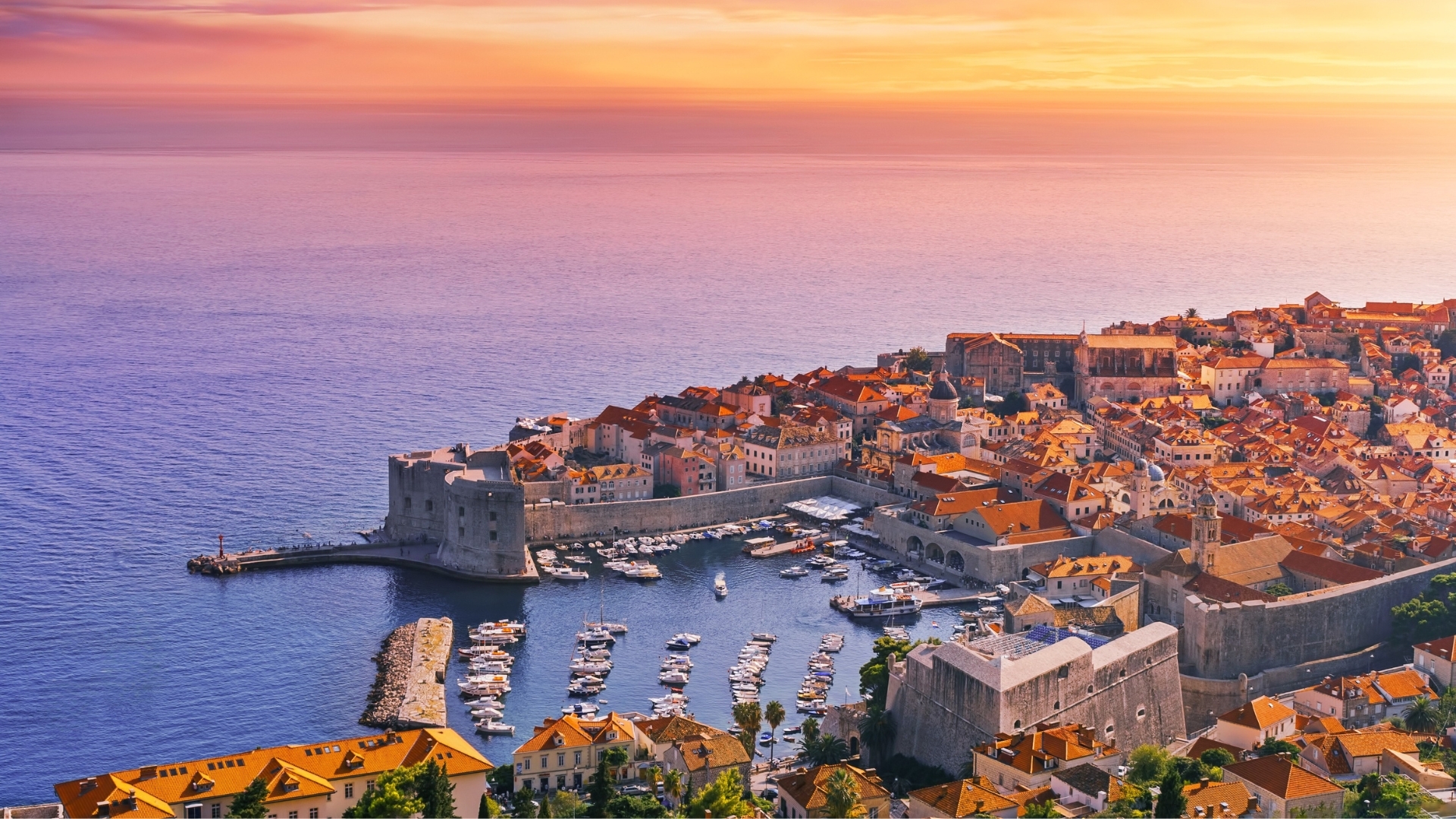 The image depicts a stunning sunset over the coastal city of Dubrovnik, Croatia. The scene features a vibrant sky with hues of orange and purple, casting a warm glow over the historic buildings with red rooftops and the calm sea. The harbor is filled with boats, and ancient city walls can be seen along the shoreline, highlighting the city's rich history and picturesque landscape.