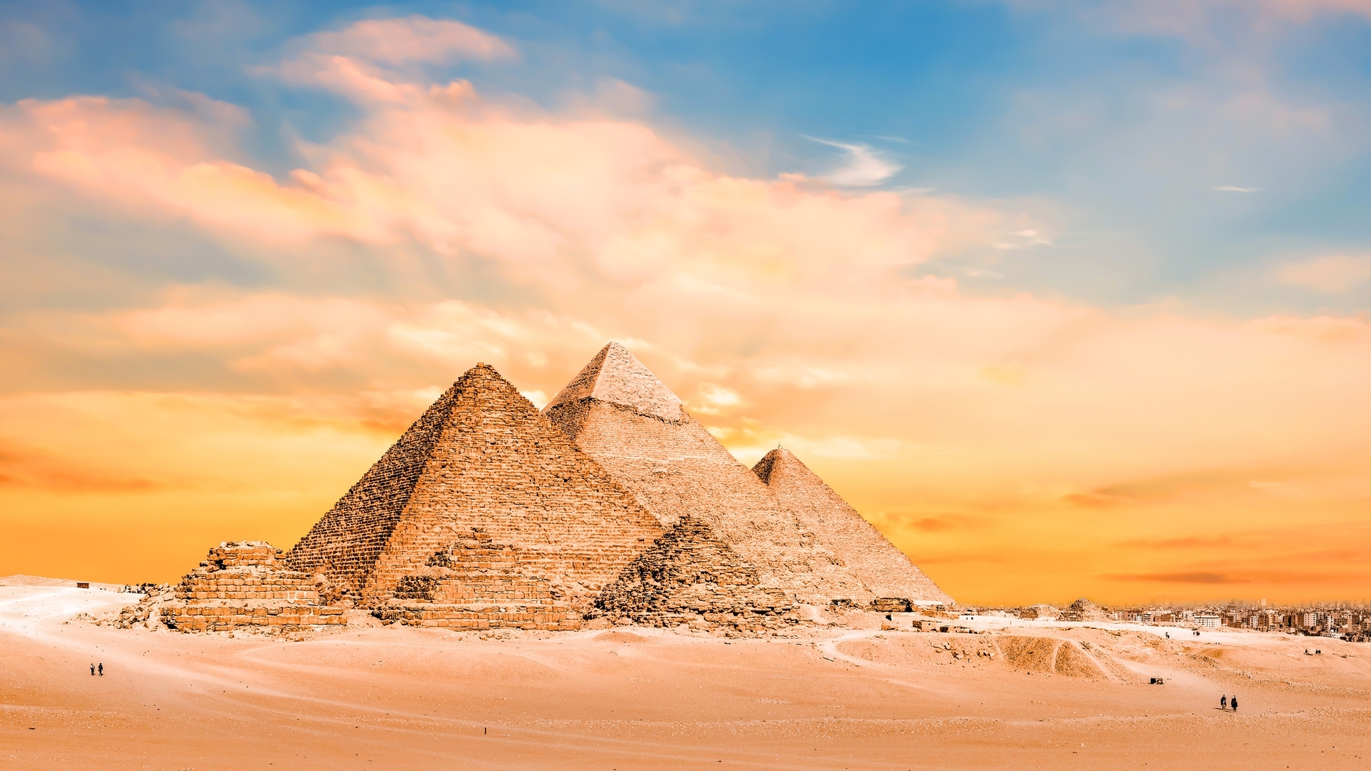 The image depicts the Great Pyramids of Giza in Egypt during sunset, showcasing their majestic structures against a vibrant sky filled with soft clouds and warm colors. The pyramids, made of large stone blocks, stand prominently in the desert landscape, with a few small figures visible in the foreground, emphasizing their grand scale.