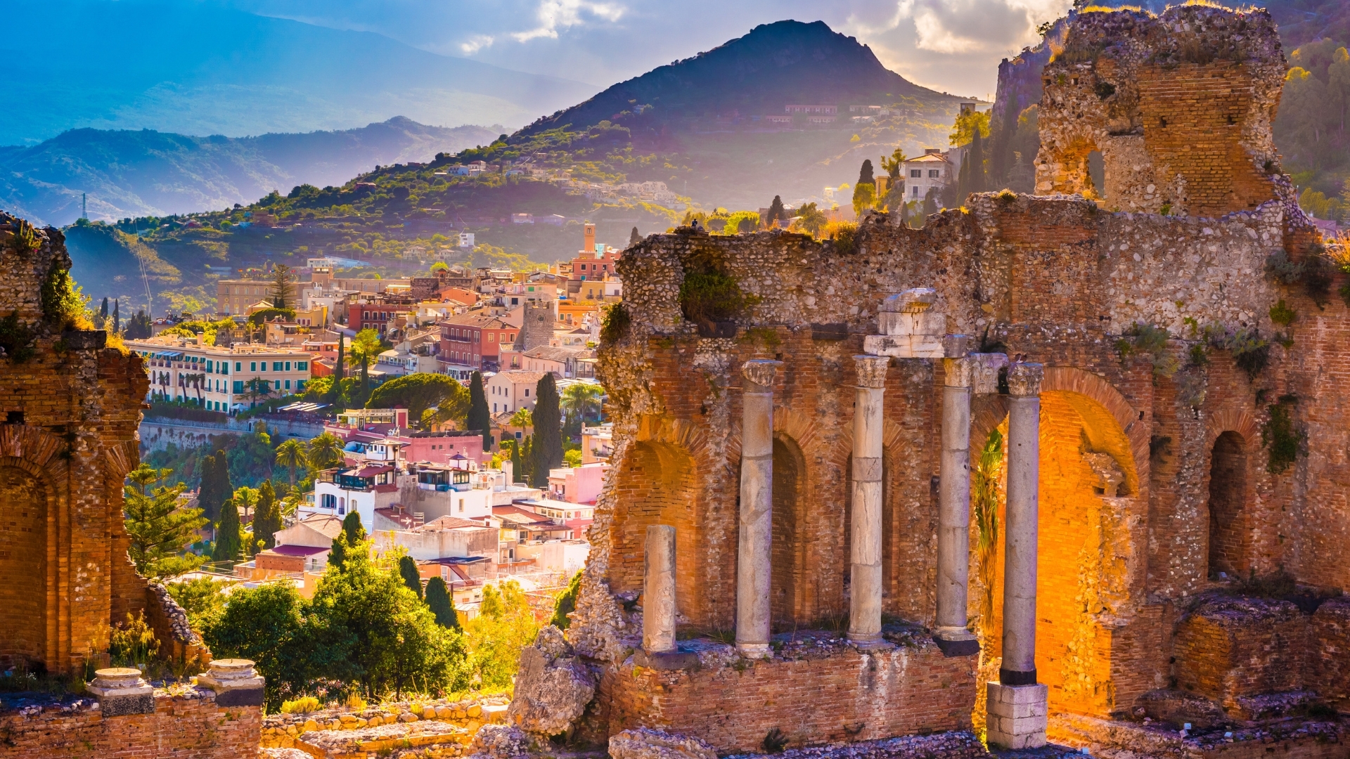 The image depicts the ruins of the ancient Taormina Theater, set against a backdrop of lush green mountains and a colorful town. The theater's stone structures, including columns and arches, are illuminated by the warm glow of sunset, creating a picturesque scene that highlights the historical significance and beauty of the location.