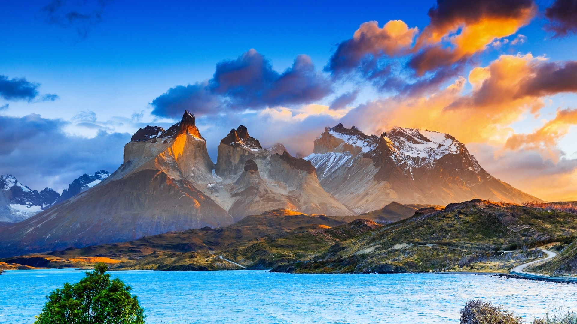 The image depicts the stunning landscape of Torres Del Paine National Park in Chile, showcasing dramatic mountains with snow-capped peaks, a vibrant blue lake in the foreground, and a colorful sky filled with clouds at sunset.