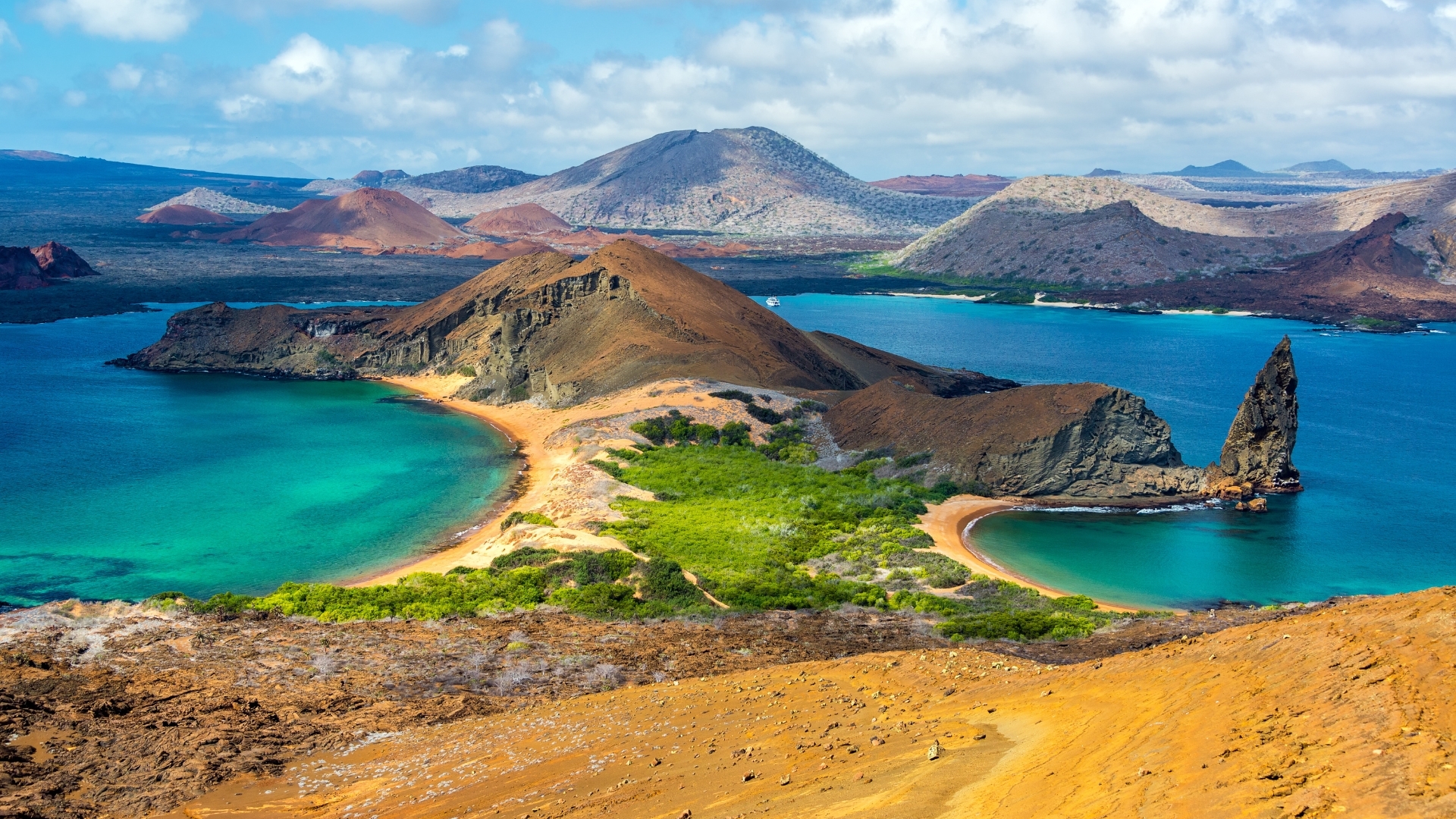 The image shows a scenic view of Bartolome Island, featuring a sandy beach, lush green vegetation, and a prominent rock formation jutting out of the water. The landscape includes turquoise waters and a backdrop of volcanic hills and mountains under a partly cloudy sky.