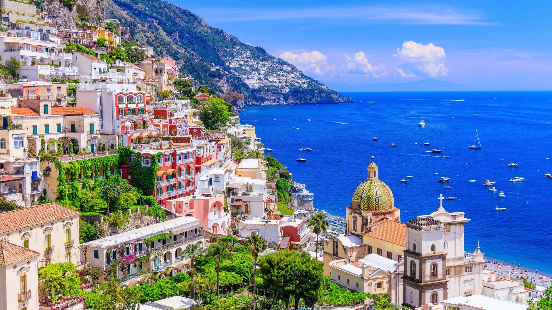 The image depicts a vibrant view of Positano, a picturesque town on the Amalfi Coast in Italy. Colorful buildings cascade down a steep hillside towards the blue sea, with boats dotting the water and lush greenery surrounding the structures. A prominent dome with intricate patterns is visible, adding to the town's charm.