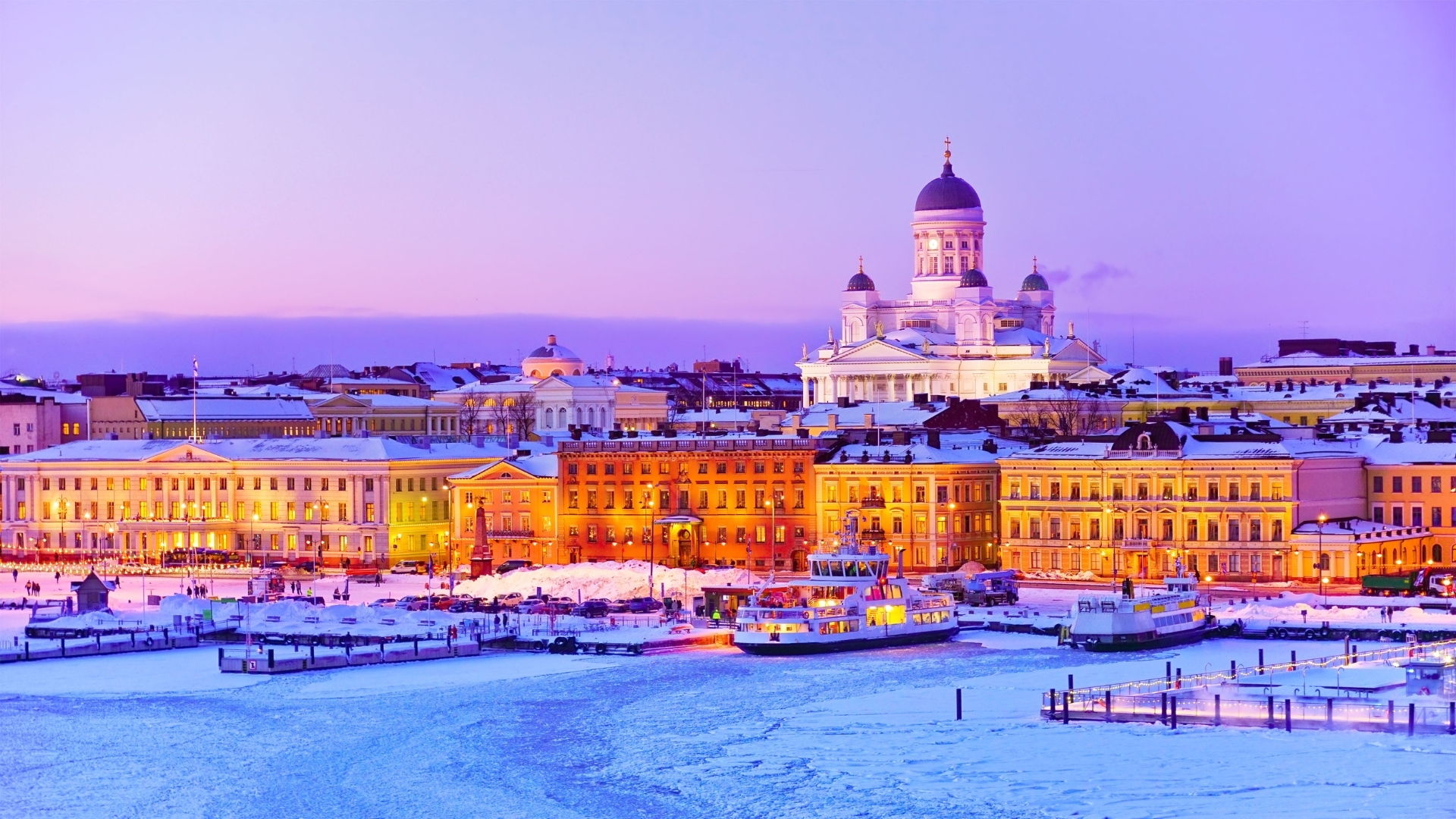 The image depicts a beautiful winter scene of Helsinki, Finland, at dusk. The harbor is icy, and the buildings along the waterfront are illuminated with warm lights, contrasting against the snowy landscape and the soft purple sky. The iconic Helsinki Cathedral is visible in the background, adding to the charm of the scene.