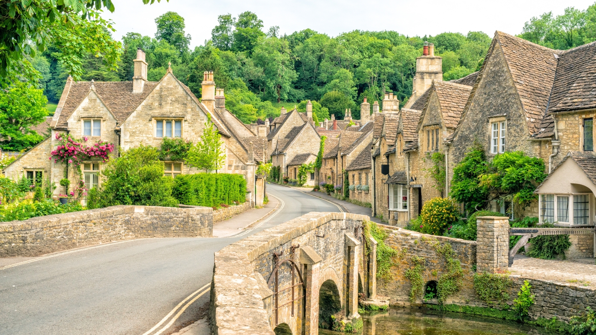 The image depicts a picturesque village scene in Castle Combe, Wiltshire, UK, featuring charming stone houses with sloping roofs, lush greenery, and a winding road that leads to a quaint stone bridge over a stream.