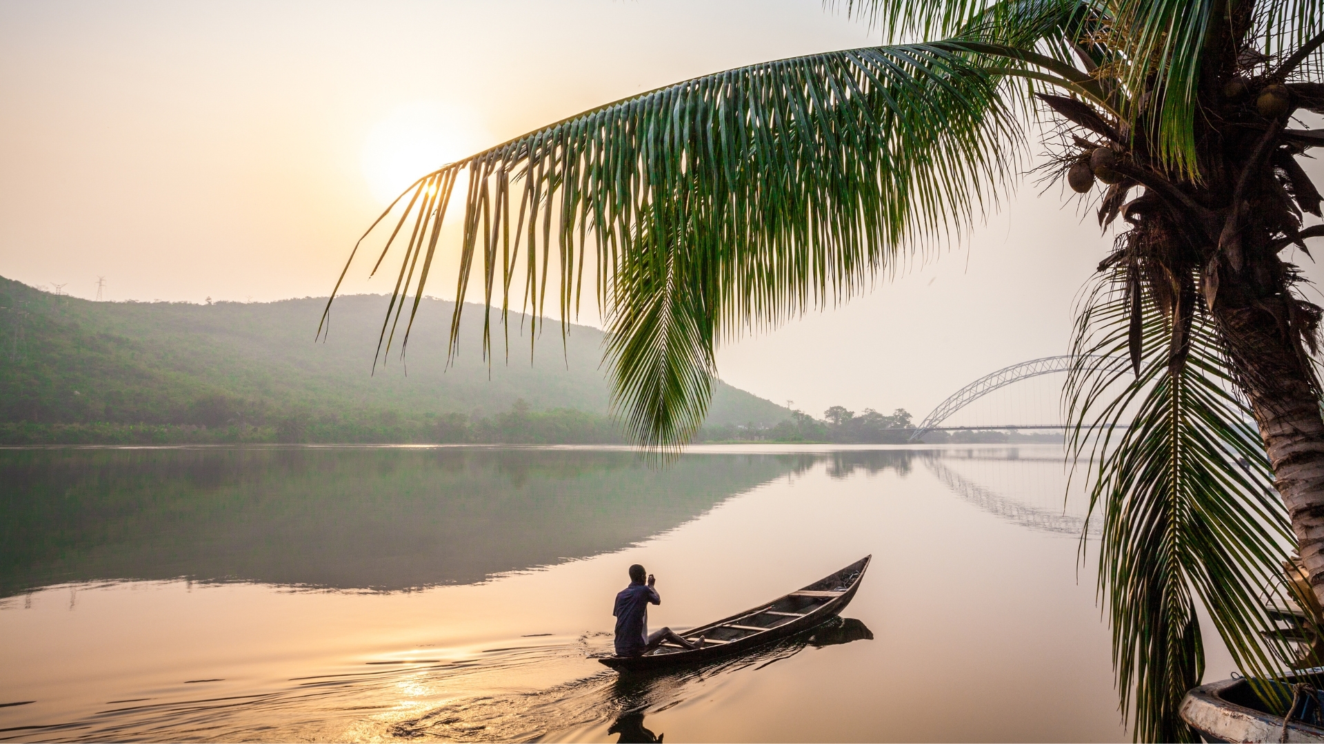 The image depicts a serene scene of the Volta River in Ghana at sunrise. A man is paddling a traditional wooden canoe on the calm water, surrounded by lush green hills and palm trees. In the background, a bridge arches over the river, reflecting the soft light of the morning sun.
